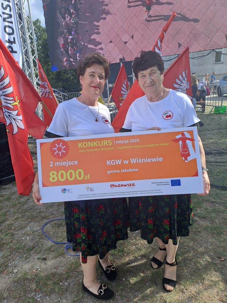 Dwie kobiety na świeżym powietrzu trzymają duży czek o wartości 8000 zł. Za nimi są czerwone flagi i duży ekran z tłumem. Ubrane są w białe koszulki i ciemne spódnice.