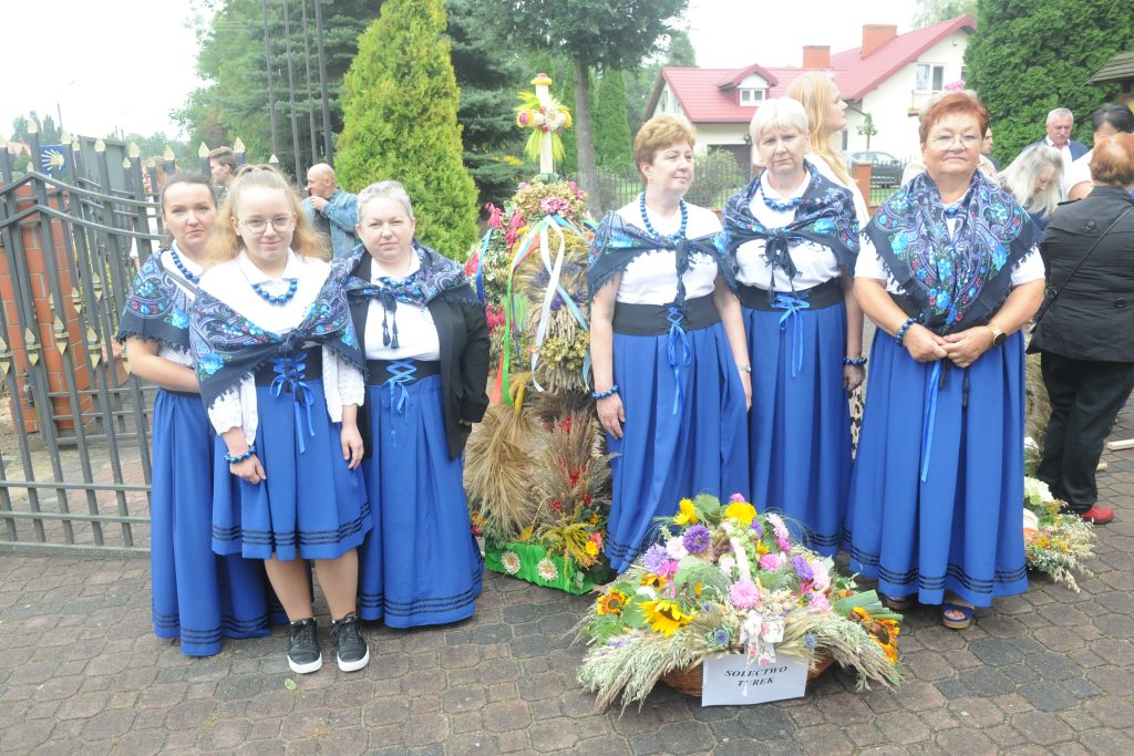 Sześć kobiet w niebieskich strojach ludowych stoi przed ozdobnym wieńcem z kwiatami. Tło stanowi ceglany chodnik i zieleń. Na ziemi leży kosz z kolorowymi kwiatami.