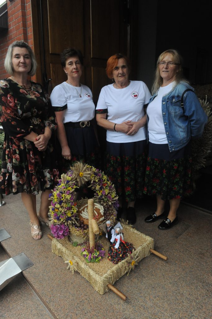 Cztery kobiety stoją przy dekoracji z zboża i kwiatów. Mają na sobie kwieciste ubrania. Tło stanowią drewniane drzwi.