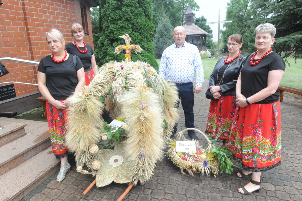 Grupa pięciu osób w ludowych strojach stoi przy dożynkowym wieńcu z kłosów zbóż. Tło stanowi ceglany budynek i zielone drzewa. Dekoracyjny wieniec zdobią kwiaty i krzyż.