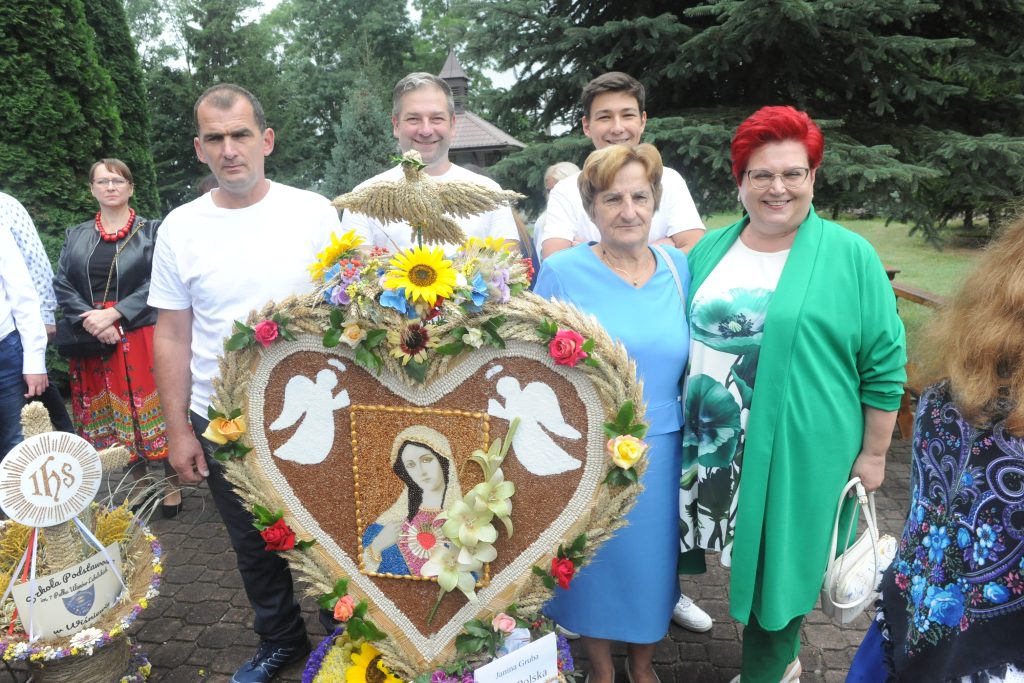 Grupa ludzi stoi wokół kolorowej ozdoby z portretem Maryi i aniołami na tle zielonych drzew. Ozdoba jest ozdobiona kwiatami. Osoby mają na sobie kolorowe stroje ludowe.