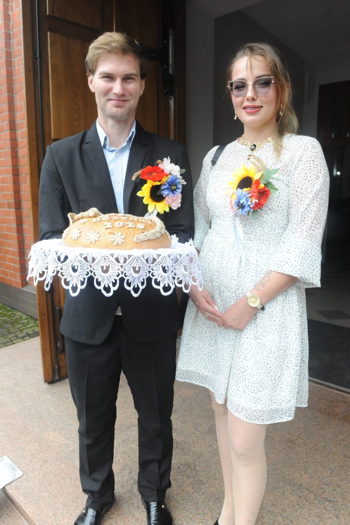 Para w eleganckich strojach trzyma chleb z dekoracjami. Kobieta w białej sukience z kropkami, mężczyzna w czarnym garniturze. Jasne tło z drewnianymi drzwiami.