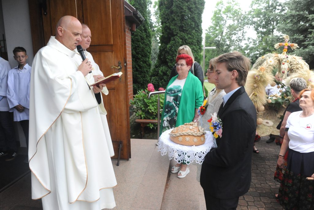 Kapłan w białej szacie czyta z księgi przed kościołem. Kobieta i mężczyzna z chlebem stoją na tle zieleni. Ozdobny wieniec zbożowy w tle.