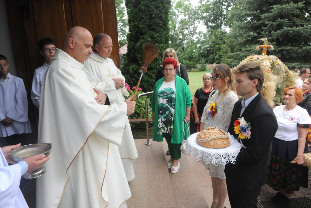 Kapłani w białych szatach błogosławią chleb w trakcie ceremonii. W tle drzewa i osoby w kolorowych strojach. Mężczyzna i kobieta trzymają chleb ozdobiony kwiatami.