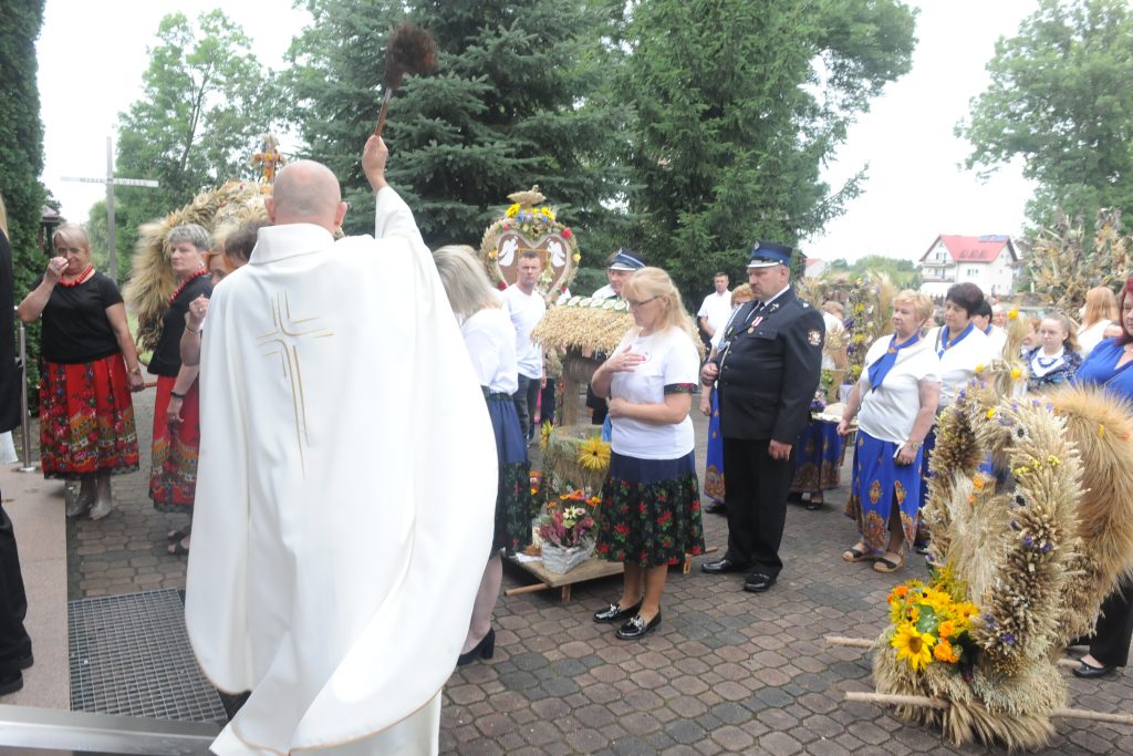 Kapłan w białej szacie błogosławi ludzi na uroczystości dożynkowej. Tłum ubrany w kolorowe stroje ludowe, w tle zieleń drzew. Kilka wieńców zbożowych zdobi miejsce.
