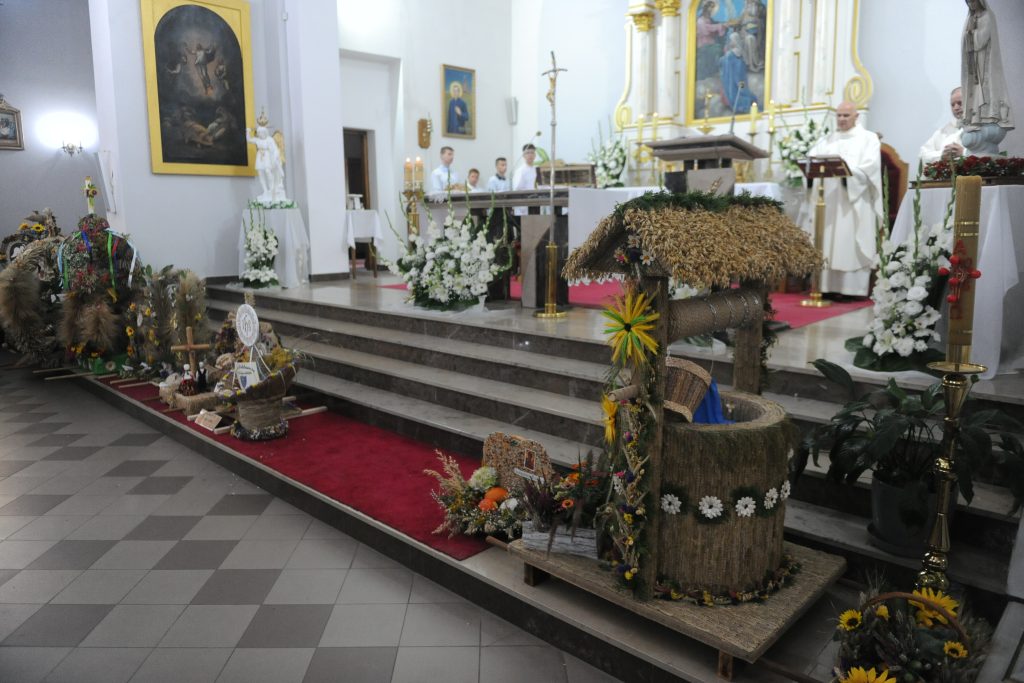 Wnętrze kościoła z ołtarzem udekorowanym białymi kwiatami. W centrum figura pokryta chlebem. Tło białe, bogate dekoracje.