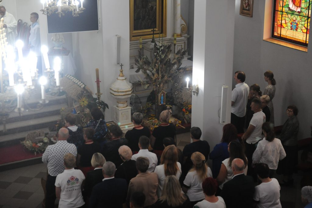 Ludzie stoją w kościele podczas ceremonii religijnej. Wnętrze jest jasne, z witrażem i dekoracjami z kwiatów i zboża. Dominuje kolor biały, złoty i drewniane detale.