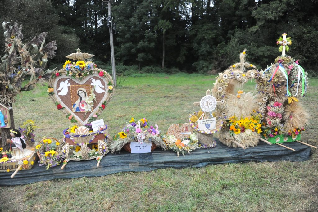 Dekoracje dożynkowe z kłosów zboża, ozdobione kolorowymi kwiatami, na tle zielonego trawnika i lasu.