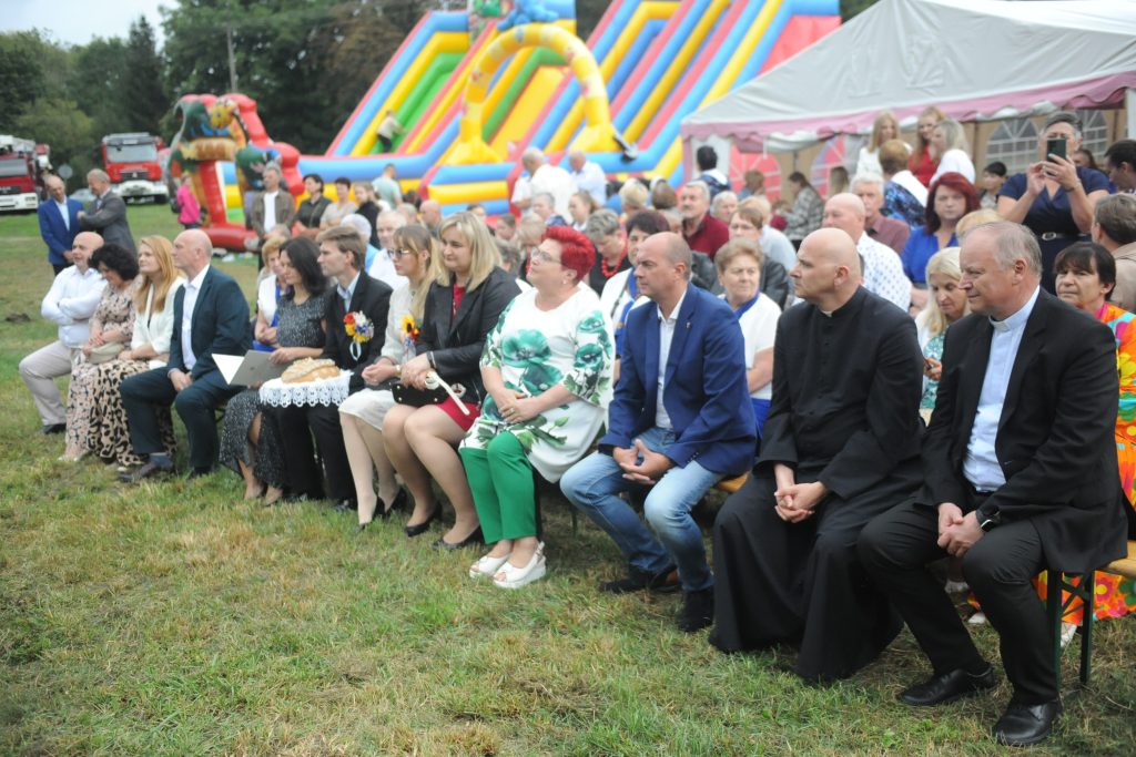 Na zdjęciu grupa osób siedzi na ławkach na trawniku. W tle widać kolorowe dmuchańce i biały namiot. Scena odbywa się na otwartym powietrzu.