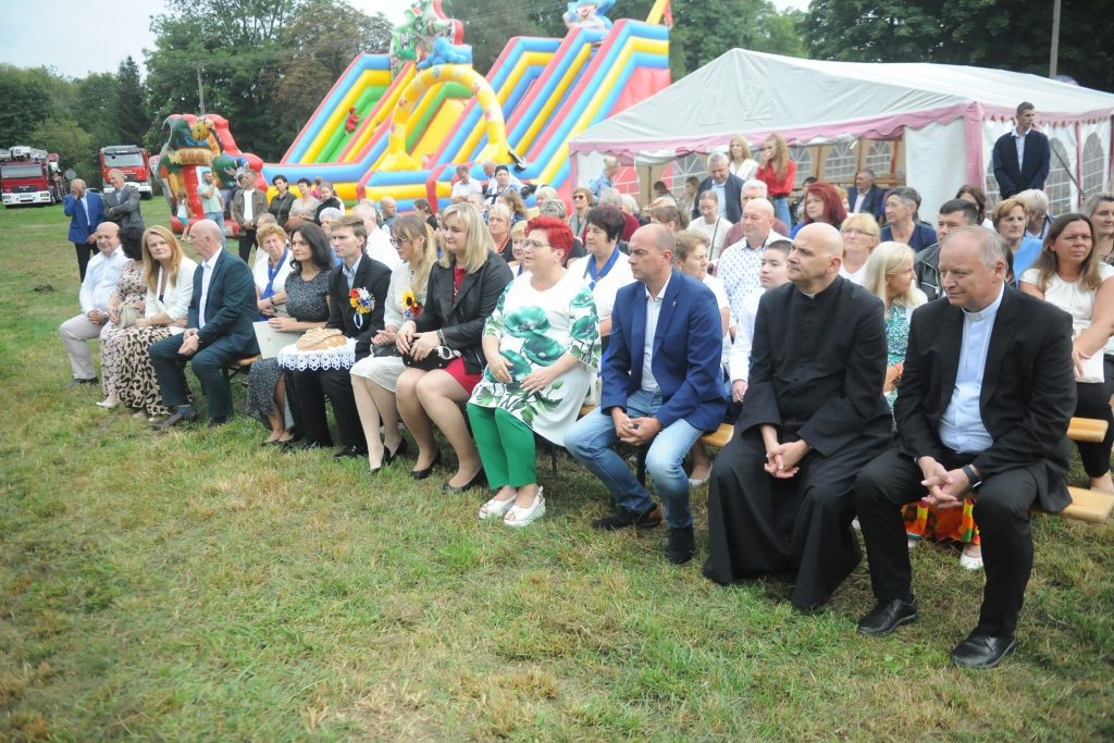 Na zielonej trawie siedzi grupa ludzi, w tle kolorowa zjeżdżalnia i biały namiot. Obok stoją czerwone wozy strażackie. Dzień jest pochmurny, otoczenie ma festynowy charakter.