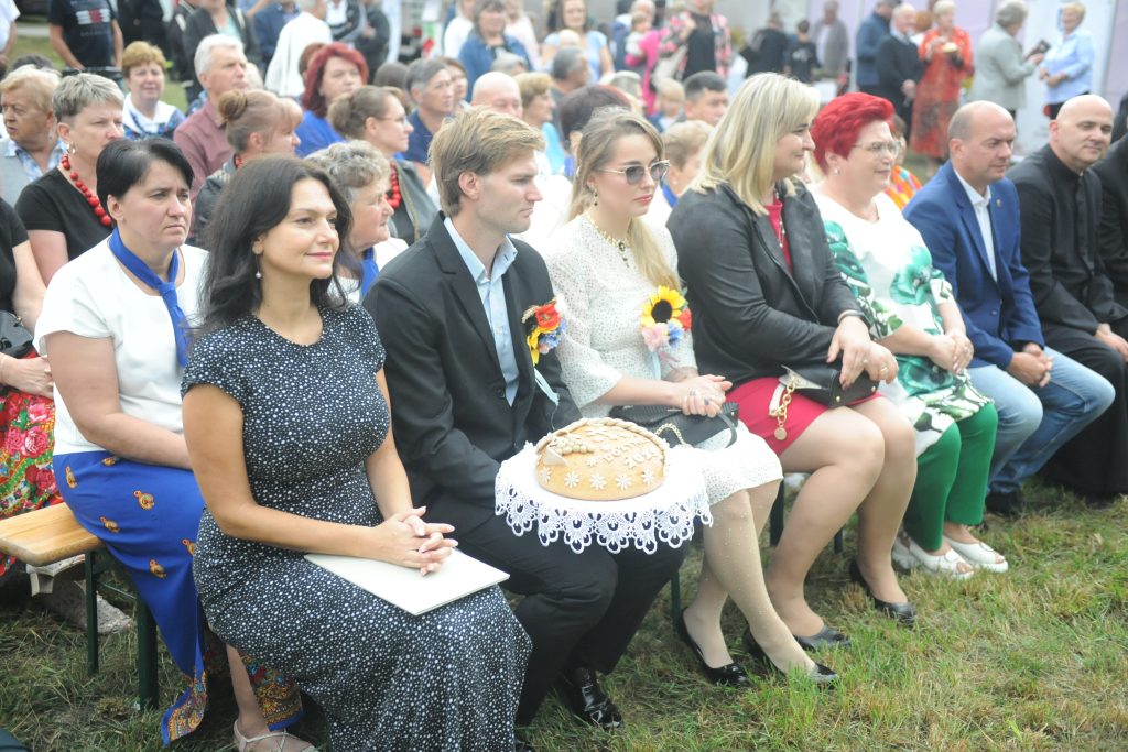 Grupa osób siedzących na ławkach na trawie podczas wydarzenia. Stroje kolorowe, z chlebem na dekoracyjnym obrusie. W tle tłum ludzi.