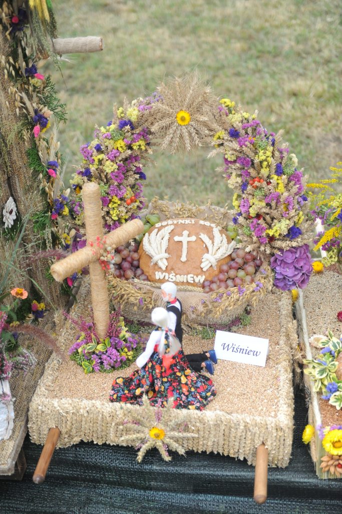 Dekoracja dożynkowa z chlebem, kolorowymi kwiatami i krzyżem. Figurka kobiety w ludowym stroju. Tło z zieloną trawą.