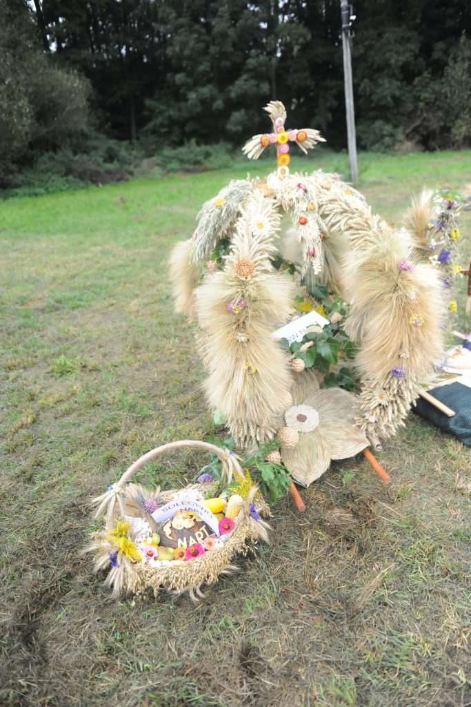 Na zdjęciu widać dożynkowy wieniec z kłosów zbóż i barwne kwiaty na trawie. W tle ciemna zieleń drzew. Obok mały koszyk z owocami i napisem.