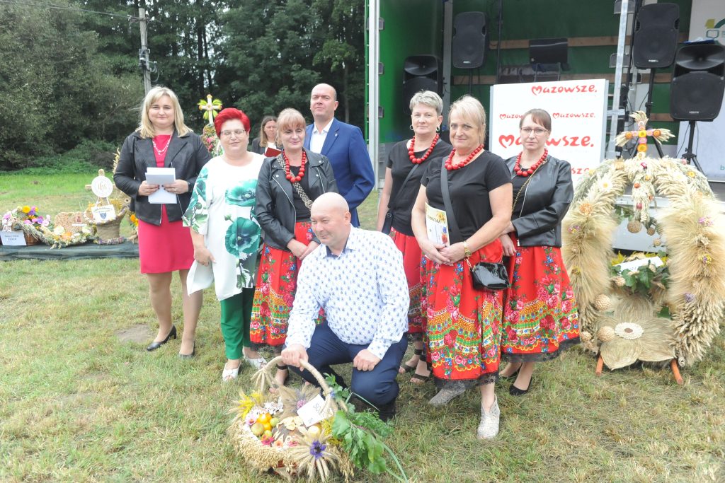 Na zdjęciu grupa ludzi stoi na trawie przed zielonymi drzewami, ubrani w kolorowe stroje ludowe z czerwonymi akcentami. W tle widać scenę z głośnikami i napisem Mazowsze. Z przodu znajduje się kosz z żniwnymi dekoracj