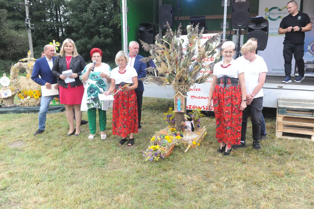 Grupa osób stoi na trawie przy scenie na tle drzew. Kobiety w czerwonych spódnicach trzymają kwiaty. Widoczne siano i ozdoby.