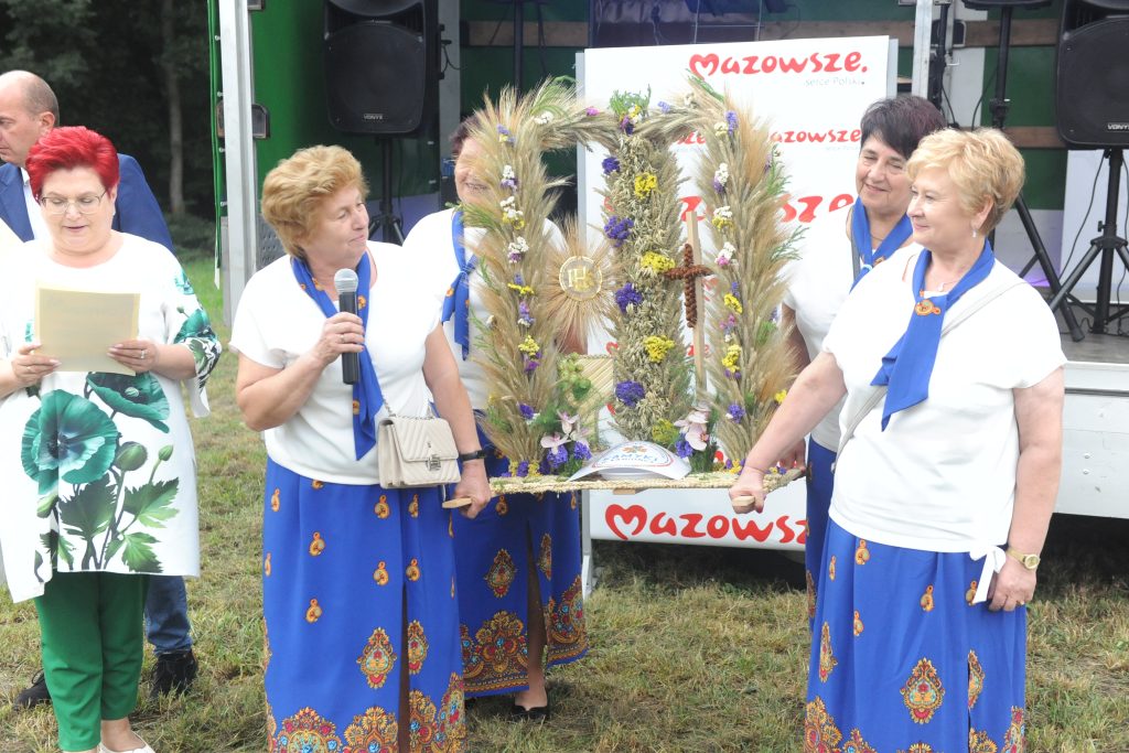 Grupa kobiet w białych bluzkach i niebieskich spódnicach trzyma ozdobny wieniec z kwiatów i kłosów. Tło to scena z napisem Mazowsze. Kobiety stoją na trawie.