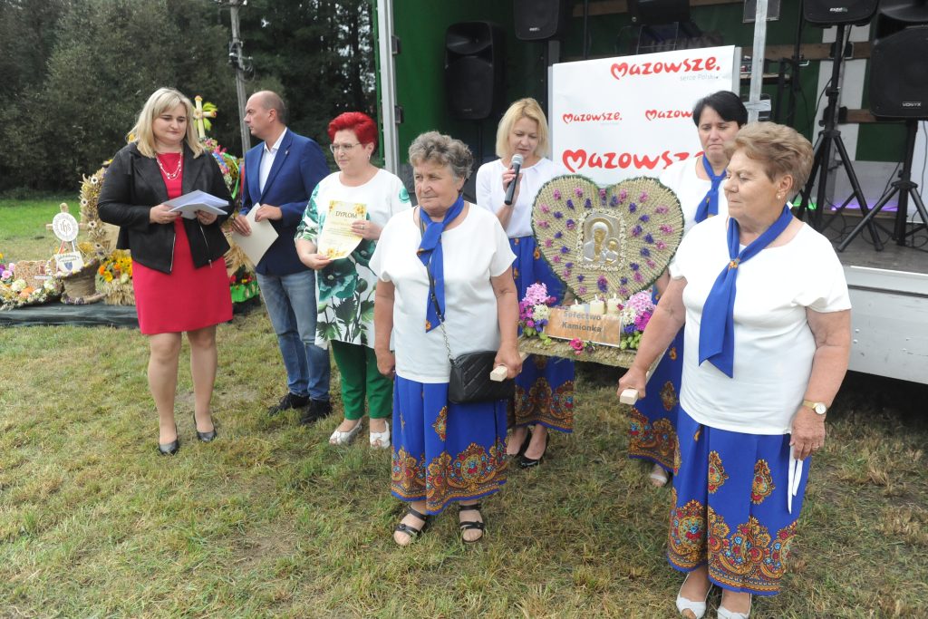 Grupa ludzi na trawie z wieńcami i certyfikatem. Kobiety w niebieskich strojach tradycyjnych z niebieskimi szarfami. W tle scena z białym banerem Mazowsze.