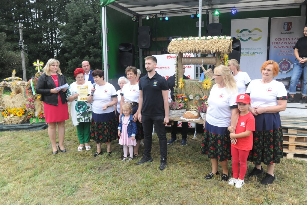 Grupa ludzi stoi na trawie, w tle scena z kwiatami i wieńcem. W centrum kobiety w ludowych strojach, jedna trzyma mikrofon. Scena jest zielona z dekoracjami, a na niej banery.