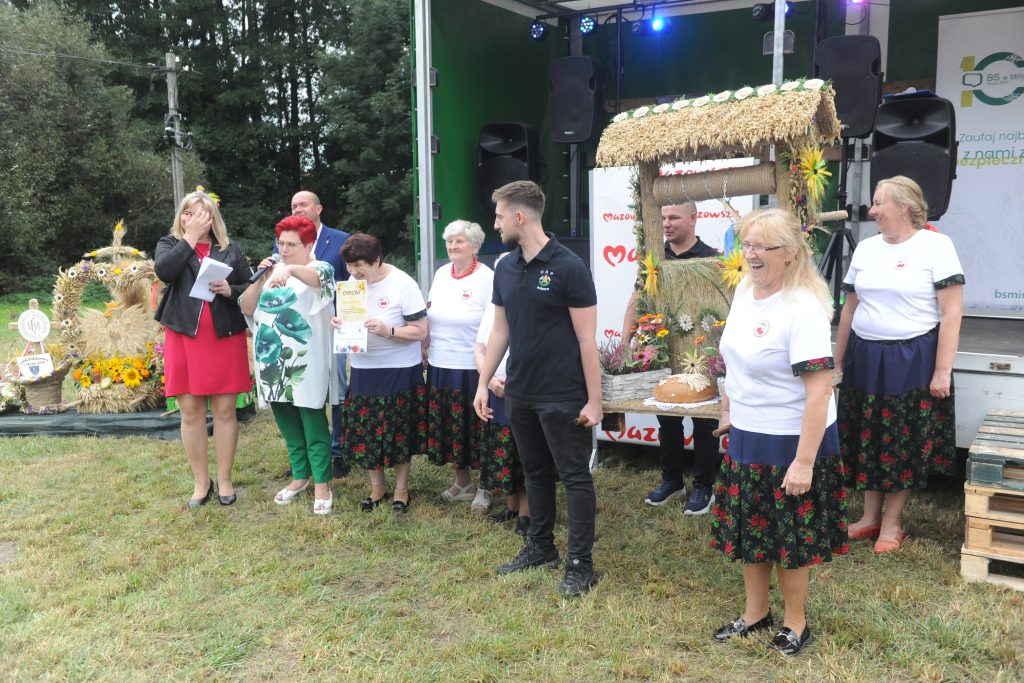 Grupa osób stoi na trawie, obok sceny z wieńcem dożynkowym. Kobiety w białych bluzkach, jedna w czerwonej sukience. Tło to drzewa i tylna część sceny.