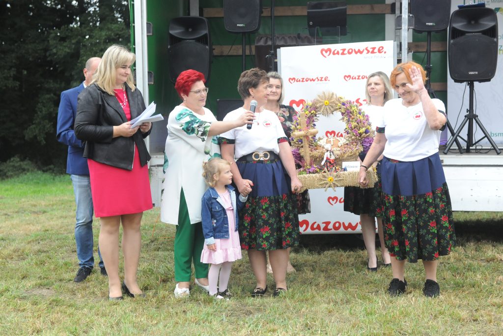 Na zdjęciu grupa ludzi na zewnątrz, na trawie, z kolorowym wieńcem. Kobiety w tradycyjnych strojach; tło z logo Mazowsze. Tło zielone, atmosfera festynu.