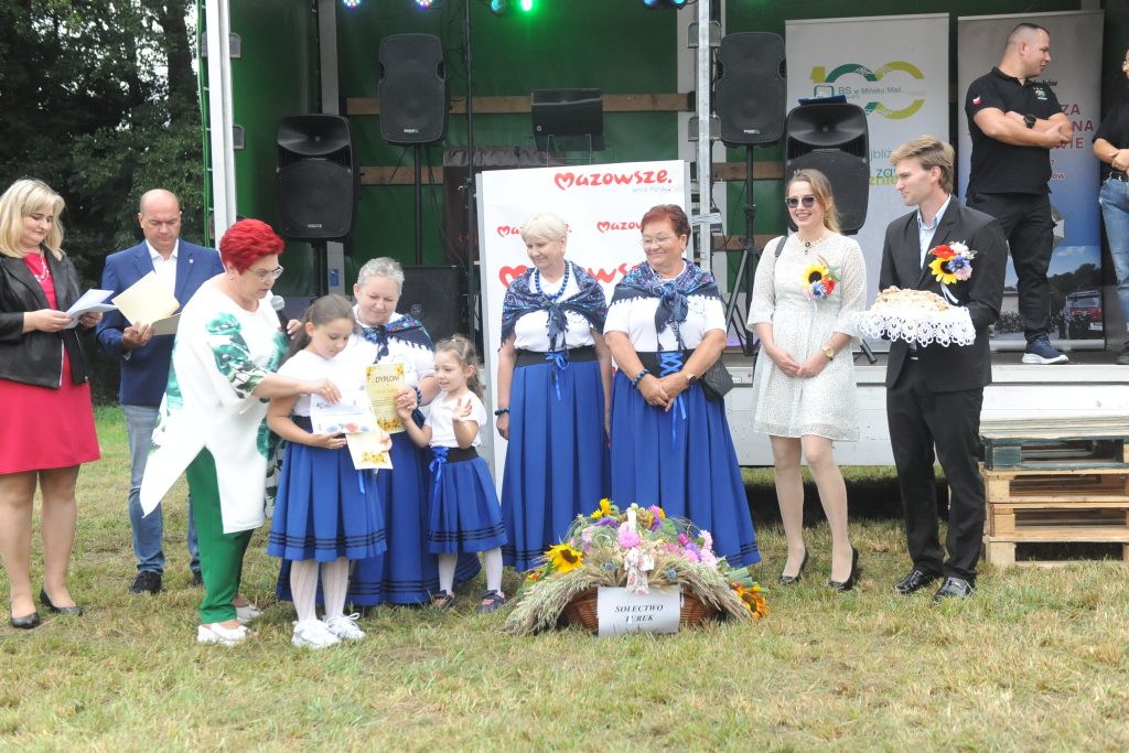 Grupa osób na scenie na zewnątrz, w tle zielone drzewa i głośniki. Kobiety w niebieskich strojach ludowych trzymają dyplomy. Na trawie kosz z kwiatami.