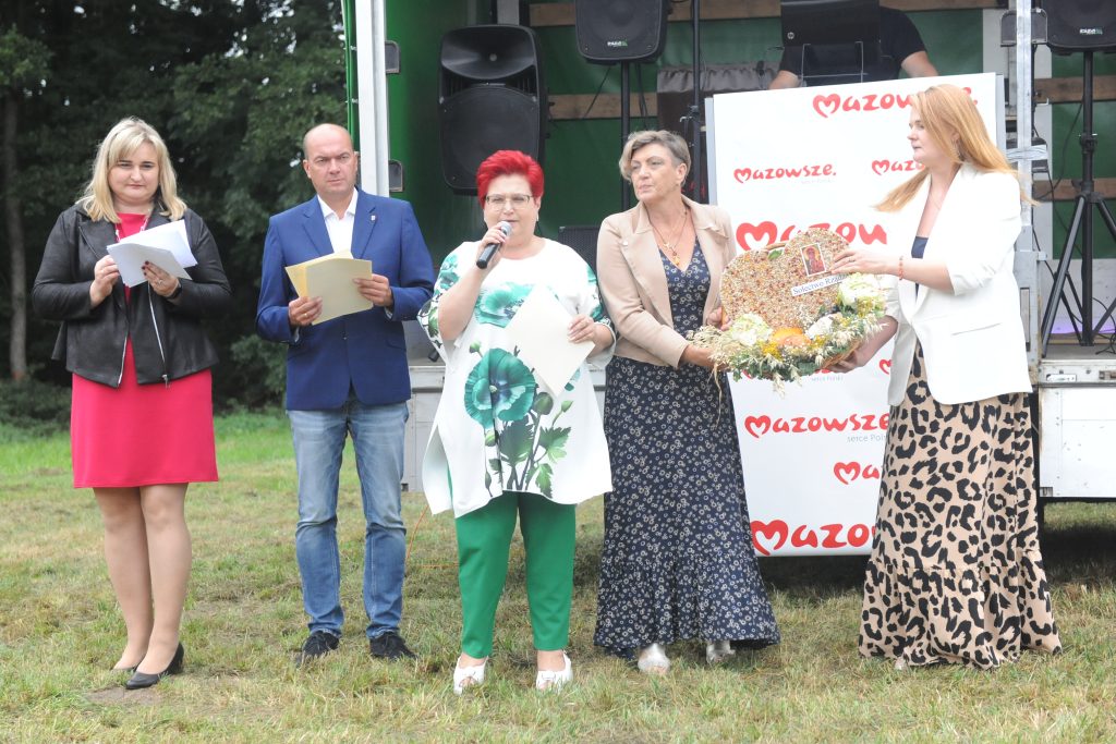 Pięć osób stoi na trawie przed sceną, na której jest napis Mazowsze. Kobieta z mikrofonem ubrana w białą bluzkę z zielonym wzorem. Obok wręczają bukiet ze zboża.