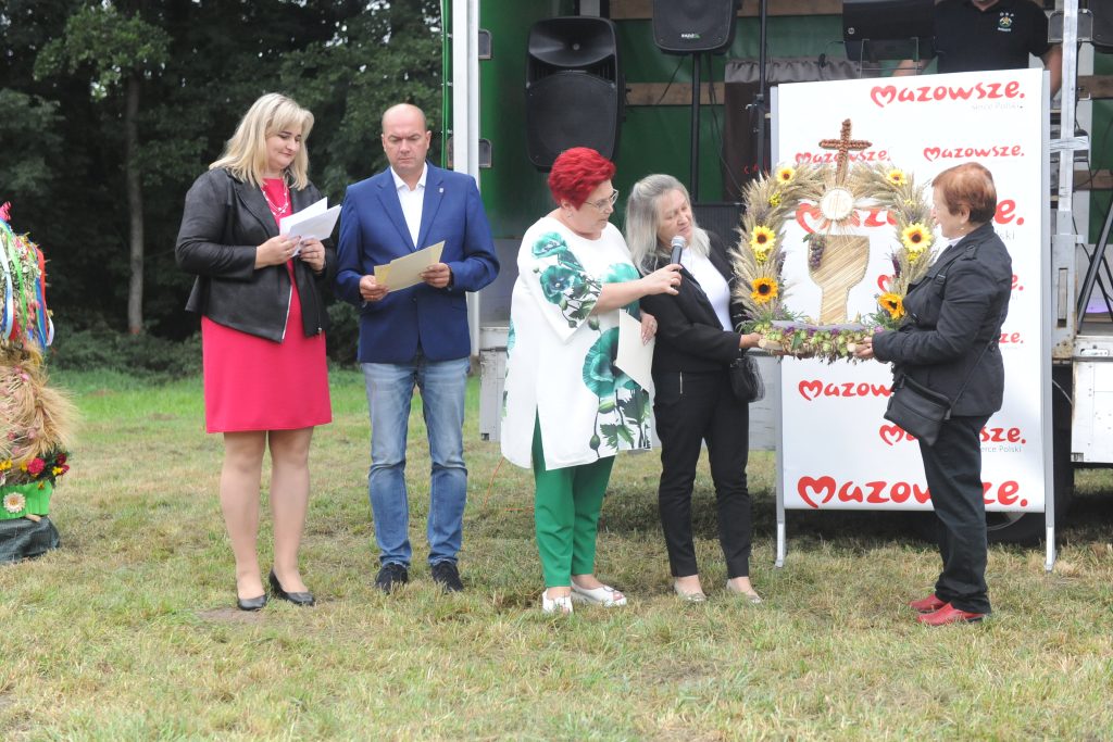 Grupa osób stoi na zielonej trawie, wręczając wieniec z kwiatów i zbóż. Na tle jest napis Mazowsze. Wszystko odbywa się na zewnątrz, przy leśnym tle.