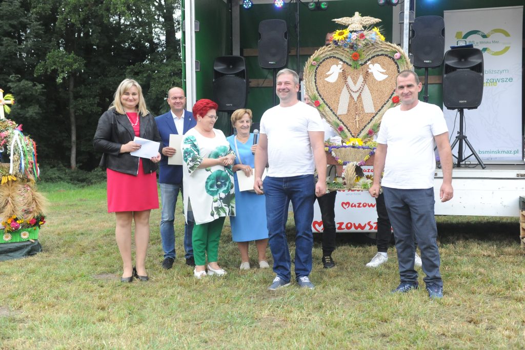 Grupa osób stoi na zielonej trawie przed sceną z dekoracją zbożową. Niebo jest zasłonięte przez drzewa. Tło sceny ma zielone i białe elementy.
