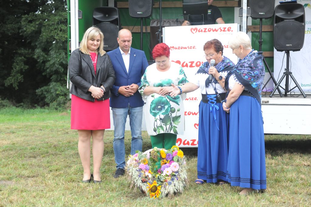 Na zdjęciu grupa osób stoi na trawie, z tyłu białe tło z napisem Mazowsze. Przed nimi leży kolorowy wieniec kwiatowy. Dwie osoby mają niebieskie stroje ludowe.