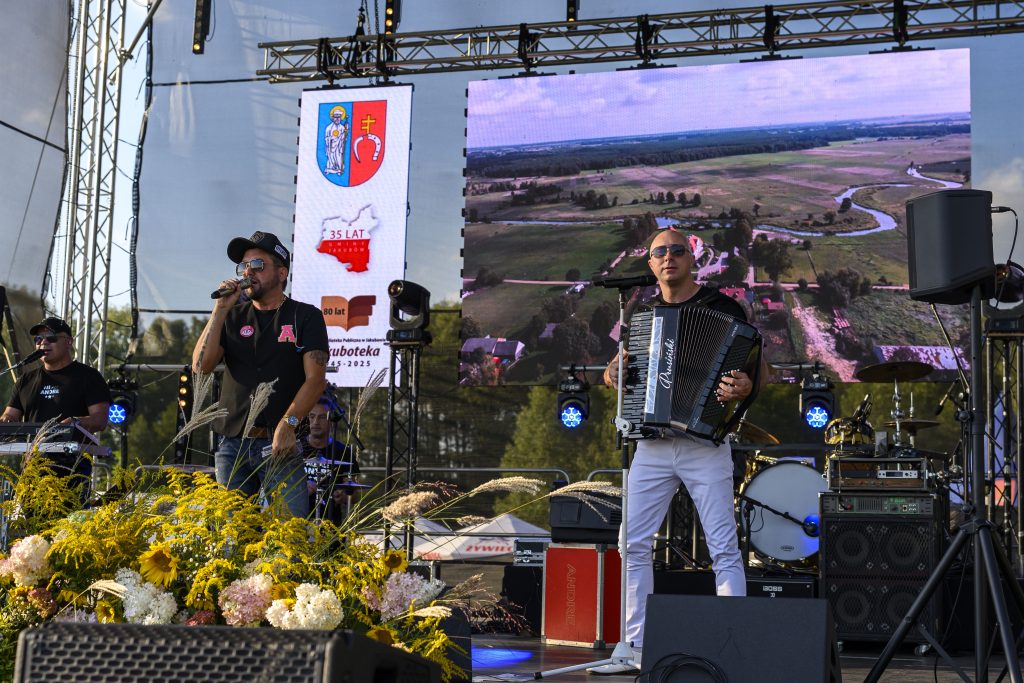 Na scenie dwóch muzyków, jeden z mikrofonem, drugi z akordeonem. Tło to duży ekran z widokiem lotniczym. Przed nimi kolorowe kwiaty.