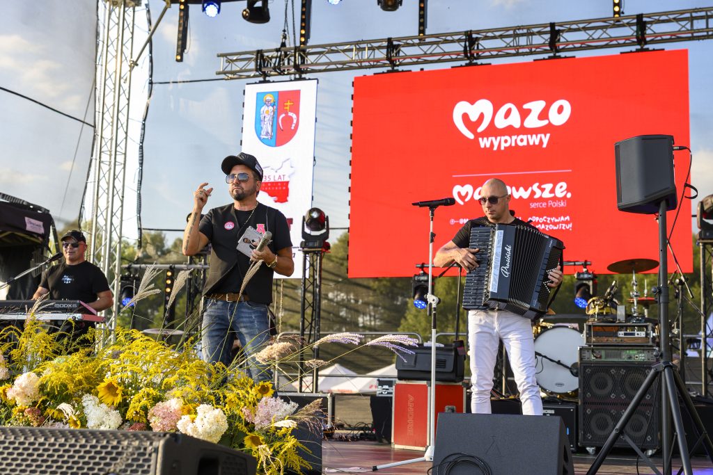 Mężczyźni na scenie z akordeonem i mikrofonem, z tyłu czerwony baner Mazo wyprawy. Przód ozdobiony żółtymi kwiatami. Tło to parkiet sceniczny i wyposażenie koncertowe.