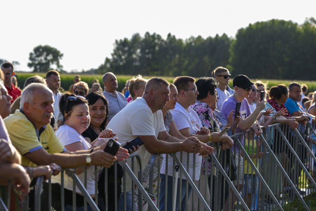 Grupa ludzi stoi za metalową barierką. Ubrani są kolorowo, patrzą w jednym kierunku. W tle widać drzewa.