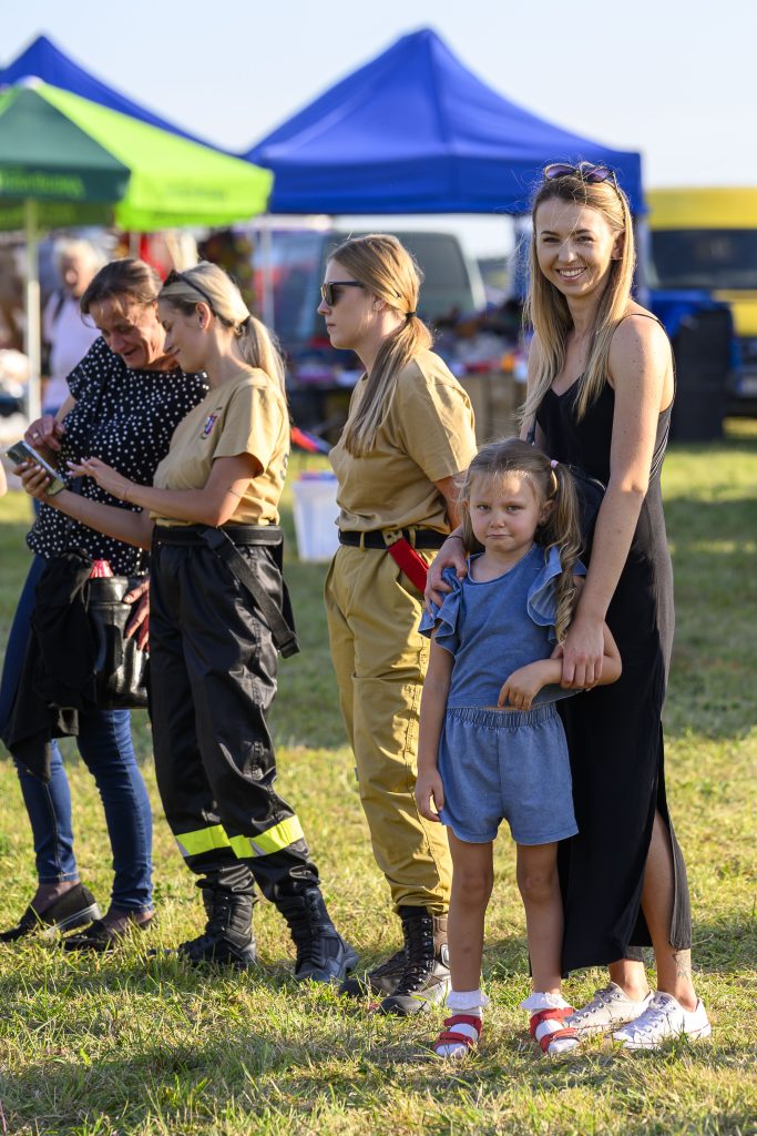 Grupa ludzi na trawie, dwie kobiety w strojach strażackich, obok niebieskie i zielone namioty. Słońce świeci intensywnie. Mała dziewczynka w niebieskim stroju stoi z kobietą.