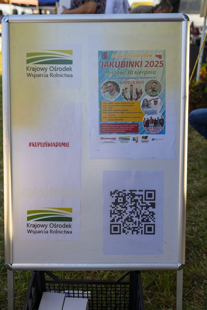 Plakat na stojaku z logotypem Krajowego Ośrodka Wsparcia Rolnictwa na białym tle. Widoczny jest kolorowy plakat zapowiadający Dni Gminy Jakubów 2025 z programem wydarzeń. Obok znajduje się QR kod i napis #KUPU