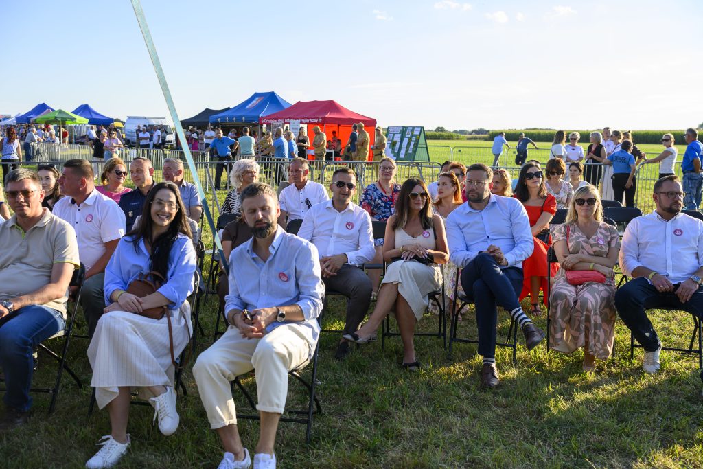 Grupa ludzi siedzi na polu w słoneczny dzień, w tle są kolorowe namioty i zielone pola uprawne. Mają na sobie letnie ubrania i okulary przeciwsłoneczne. Atmosfera pikniku lub lokalnego festynu.