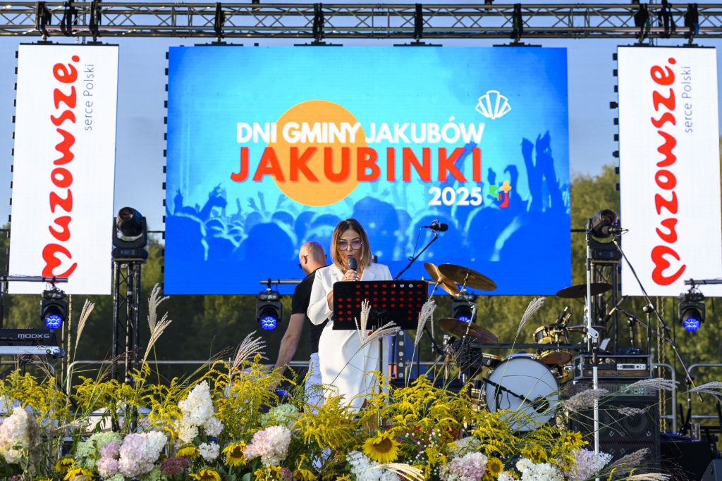 Kobieta w białym garniturze przemawia na scenie z kolorowym ekranem. Na scenie widać bębny i rośliny. Tło to zielone drzewa.