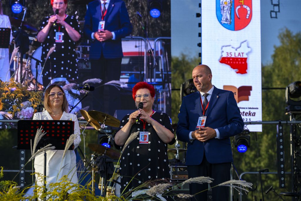 Trzy osoby stoją na scenie, przemawiają. Kobieta w czerni trzyma mikrofon, obok niej mężczyzna w garniturze. Tło jest niebieskie z żółtymi akcentami.