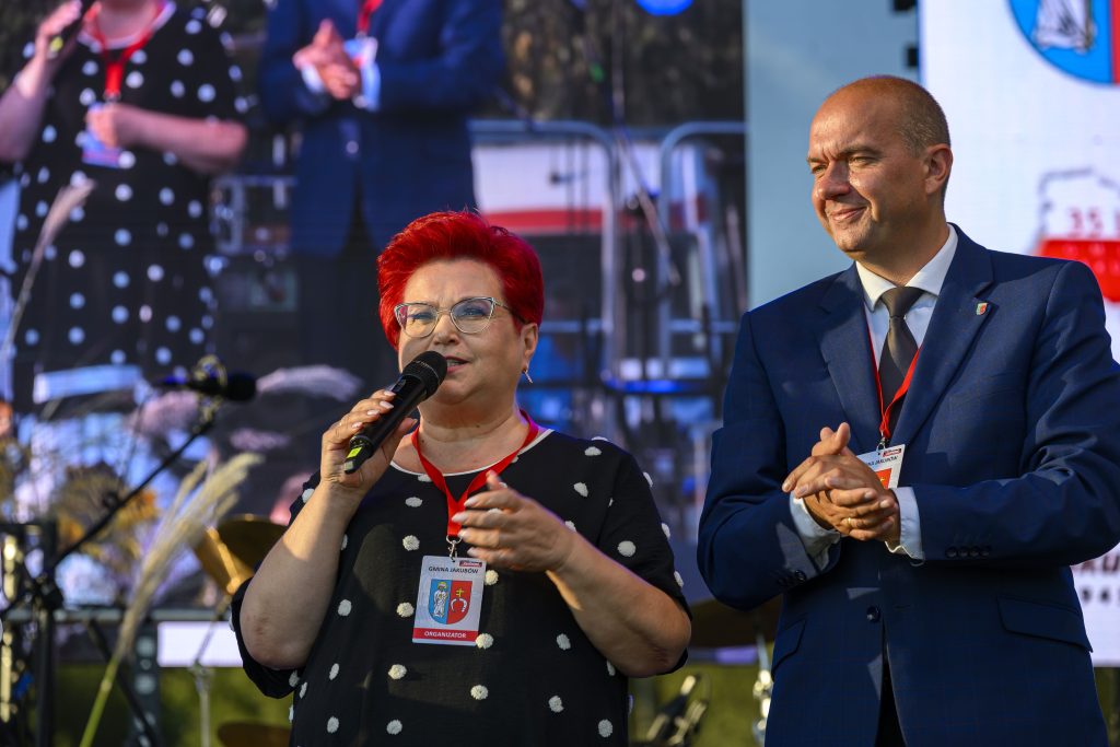 Na zdjęciu widać dwie osoby na scenie, jedna z nich z mikrofonem. Kobieta ma czerwone włosy i okulary, ubrana na czarno. Mężczyzna w garniturze stoi obok, w tle duży ekran.