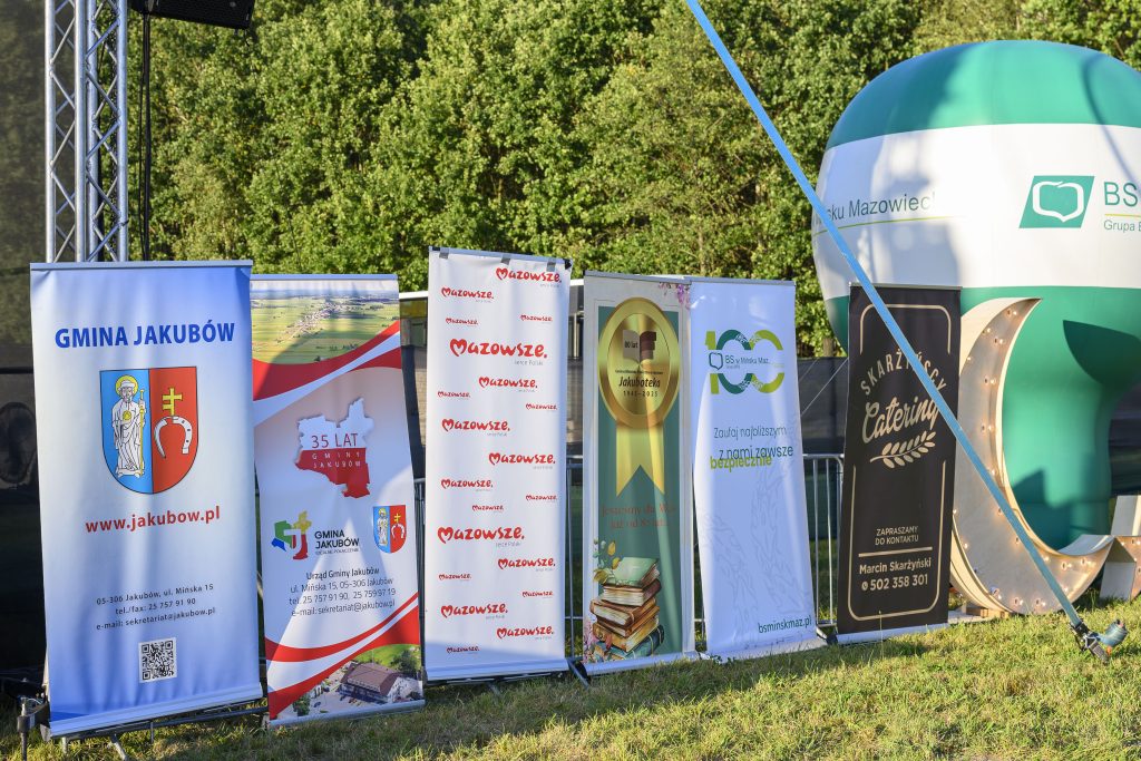 Banery na trawie promują wydarzenie w Jakubowie. Dominują kolory niebieski, zielony i biały. Tło to zielone drzewa.