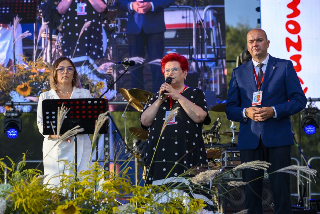 Na scenie stoją trzy osoby, jedna trzyma mikrofon. Jest kolorowy wystrój z kwiatami. Tło to duży ekran i sprzęt muzyczny.