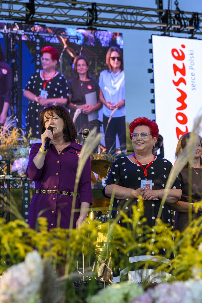 Na scenie stoi kobieta w fioletowej sukience z mikrofonem. Obok niej kobieta w kropki z czerwoną plakietką. Tło to ekran z innymi osobami i kwiaty.