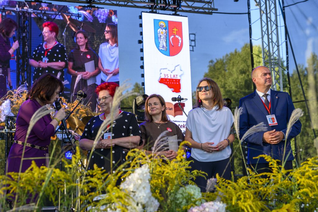 Grupa osób stoi na scenie między kwiatami, przy ekranie z herbem miasta i napisem 35 lat. Tło jest zielone i jasne.