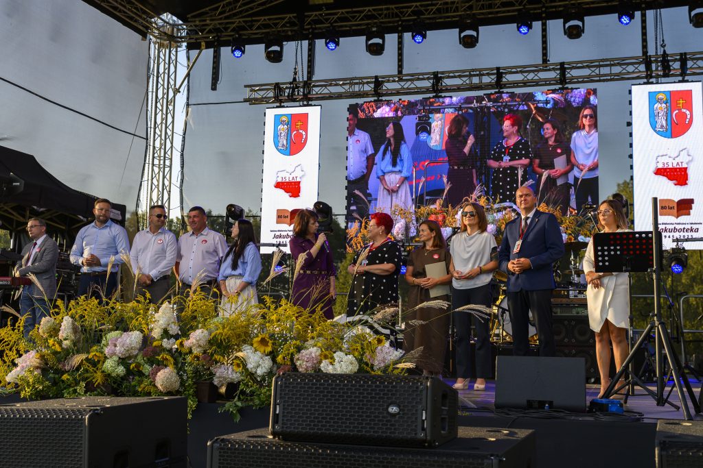 Na scenie stoi grupa ludzi obok kwiatów. W tle widać ekran z grafiką lokalną. Przewaga barw niebieskich i żółtych.