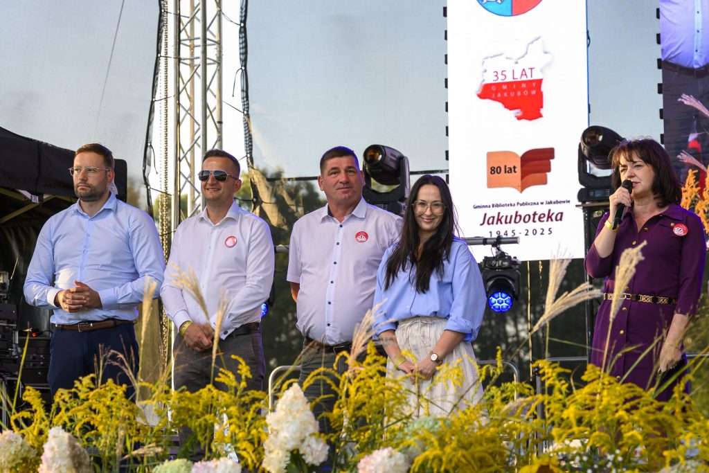 Pięć osób stoi na scenie z mikrofonem, w tle baner, a na przodzie żółte kwiaty. Ubrani są elegancko, w jasne koszule. Scena oświetlona ciepłym światłem.