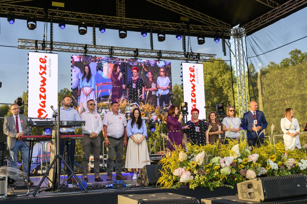 Grupa ludzi stoi na scenie z mikrofonami. Za nimi widoczny duży ekran i logo Mazowsze. Przód sceny ozdobiony kwiatami.