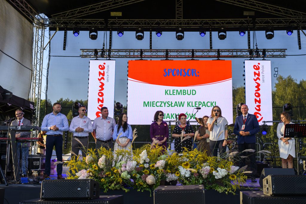 Grupa ludzi na scenie, przed kolorowym ekranem z napisem Sponsor. Dekoracje kwiatowe na przodzie, w tle drzewa.