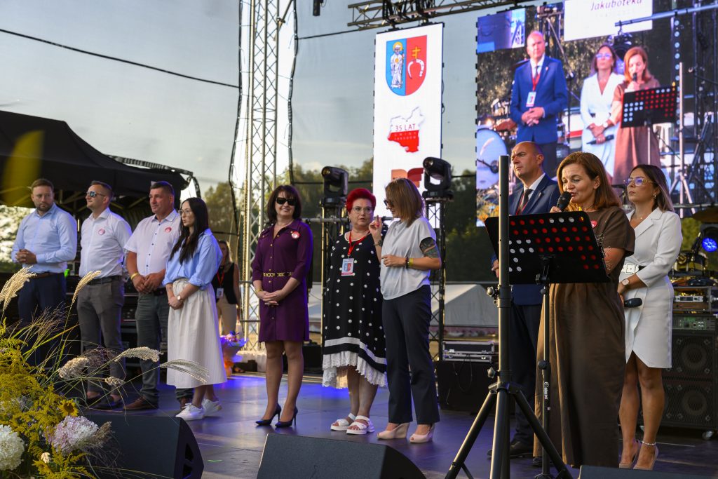 Na scenie stoi grupa ludzi w eleganckich strojach. W tle ekran z grafiką, scena jest oświetlona. Przed nimi mikrofon.