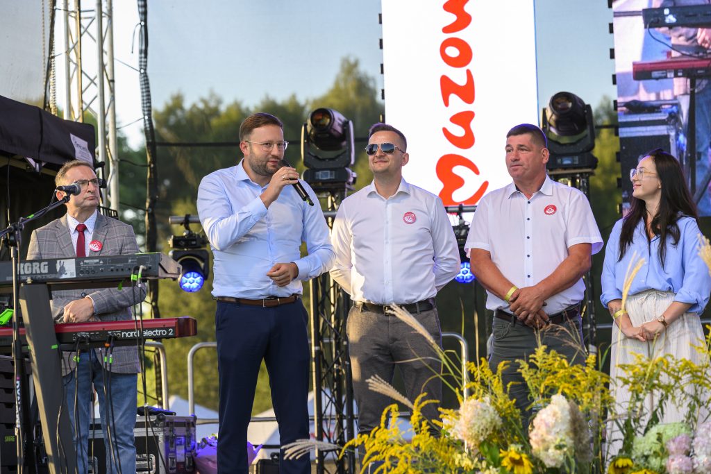 Na scenie stoi grupa ludzi w formalnych ubraniach. Jeden mężczyzna trzyma mikrofon, w tle zielone drzewa. Na pierwszym planie są kwiaty.