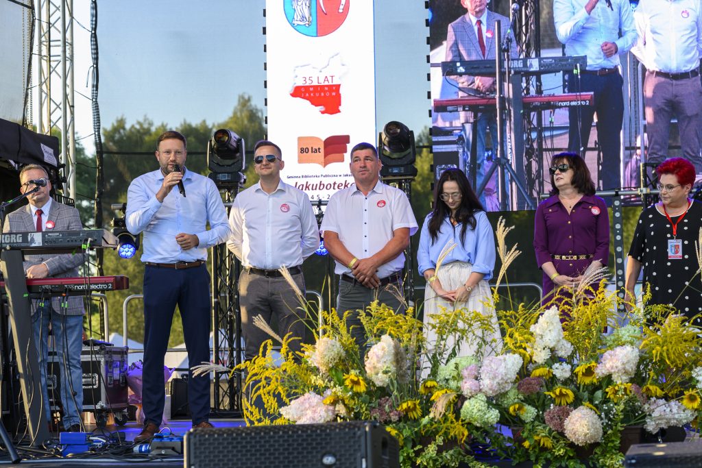 Na scenie stoi grupa osób, w tle ekran z projekcją. Dominują kolory niebieski, biały i żółty kwiatów. Za nimi zielone drzewa.
