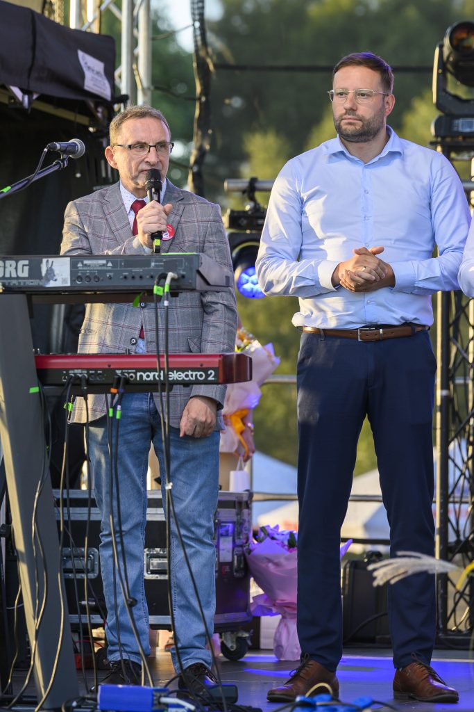 Dwóch mężczyzn na scenie, jeden mówi do mikrofonu, przed nimi syntezator. Tło to zielone drzewa i sceniczne oświetlenie.